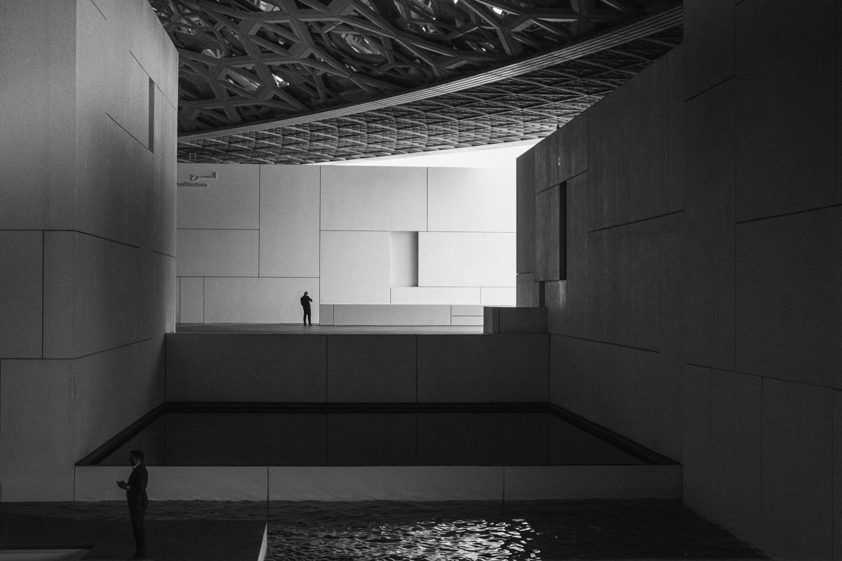 A single small figure stands in a tall pale courtyard under a patterned dome