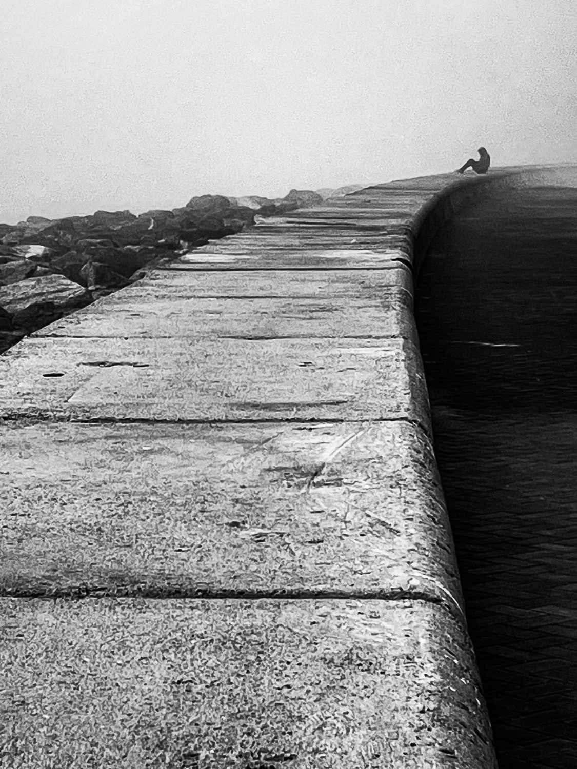 A person sits at the far end of a long stone sea wall in the mist