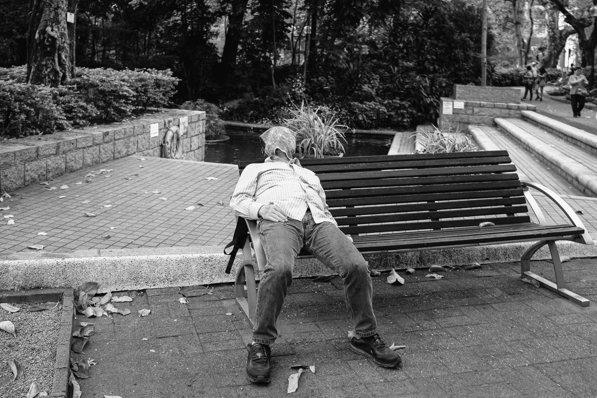 An older man in a cap sits alone on a park bench, leaning forward