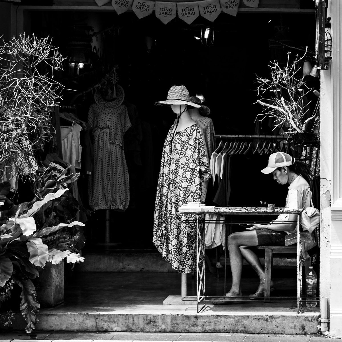 A shop assistant sits reading in a doorway between mannequins and hanging dresses
