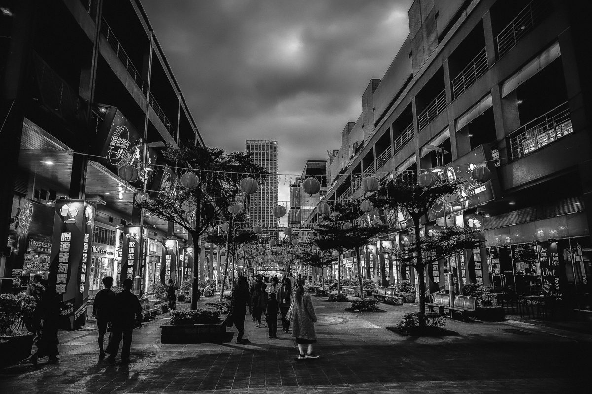 People walk through a long shopping street under a heavy sky