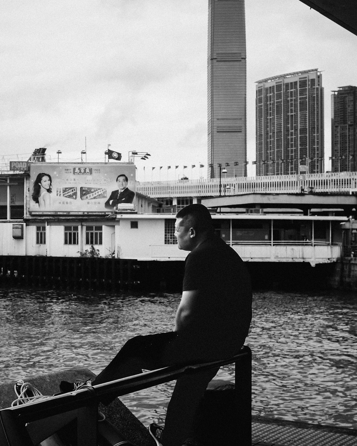 A man sits alone on a harbour railing in front of a ferry pier and towers