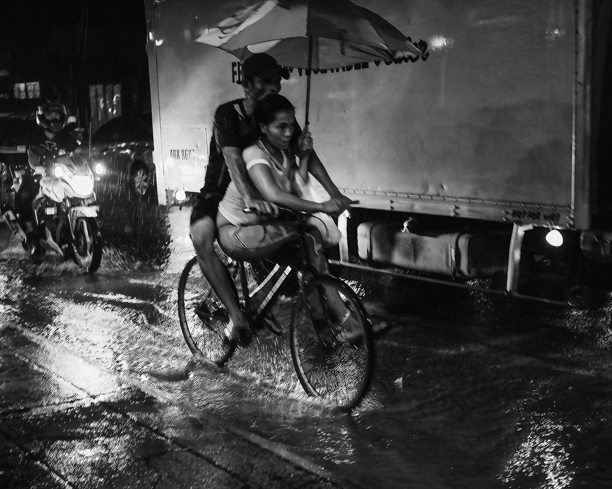 Two people share a bicycle and an umbrella through a flooded street at night