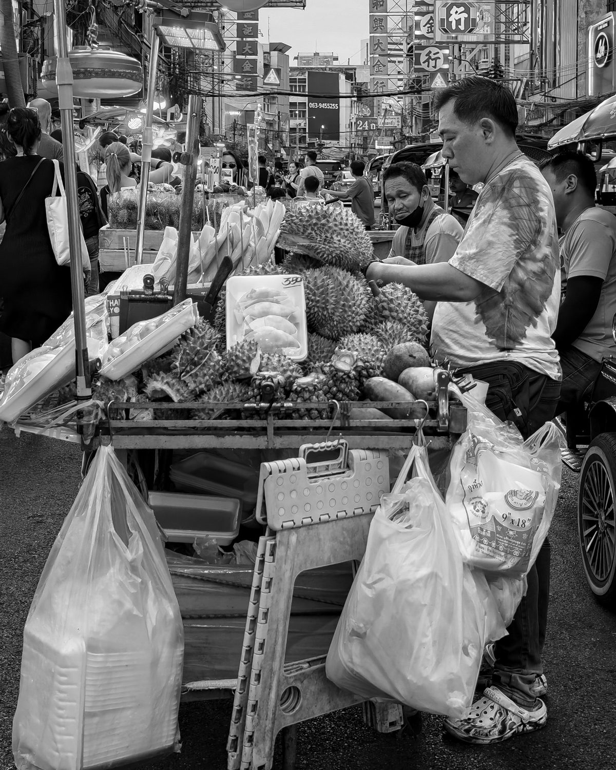A durian vendor in a flowered shirt stands behind a crowded street cart piled with fruit and plastic bags
