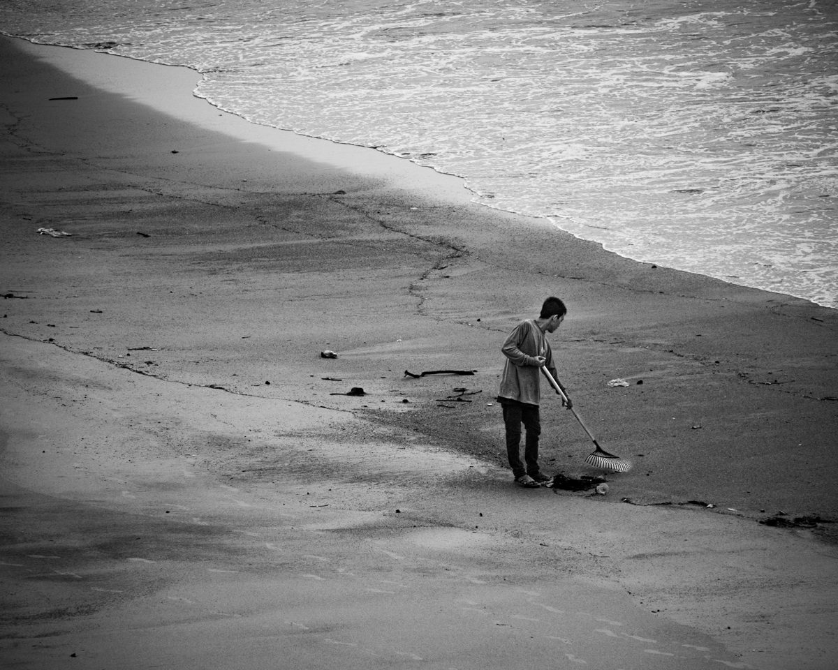 A beach worker with a rake stands alone on a stretch of wet sand beside the surf