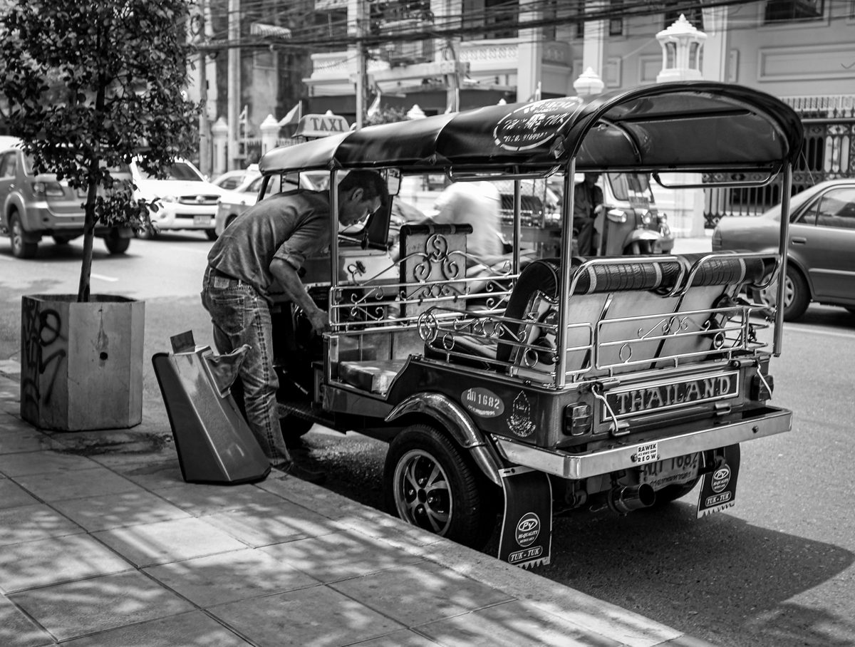 A tuk-tuk driver leans into his three-wheeled taxi parked at the kerb in dappled sunlight