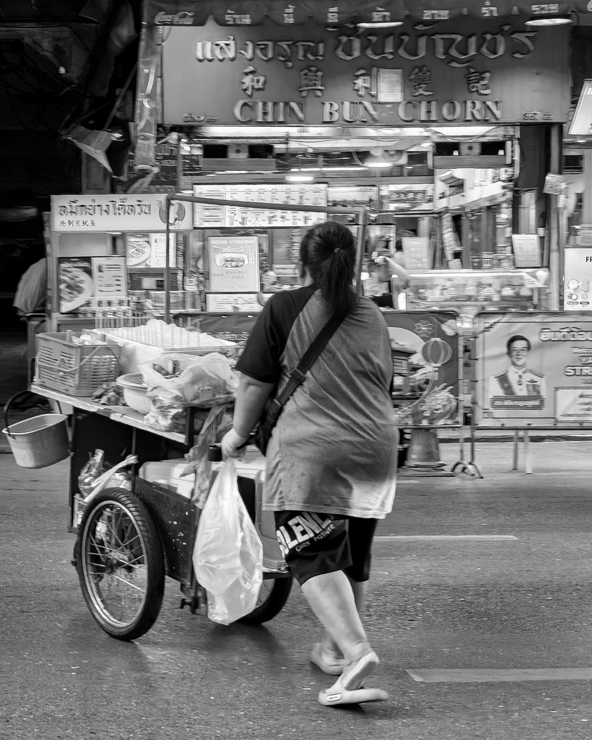 A woman in shorts walks across the street pushing a loaded handcart past a bright shopfront