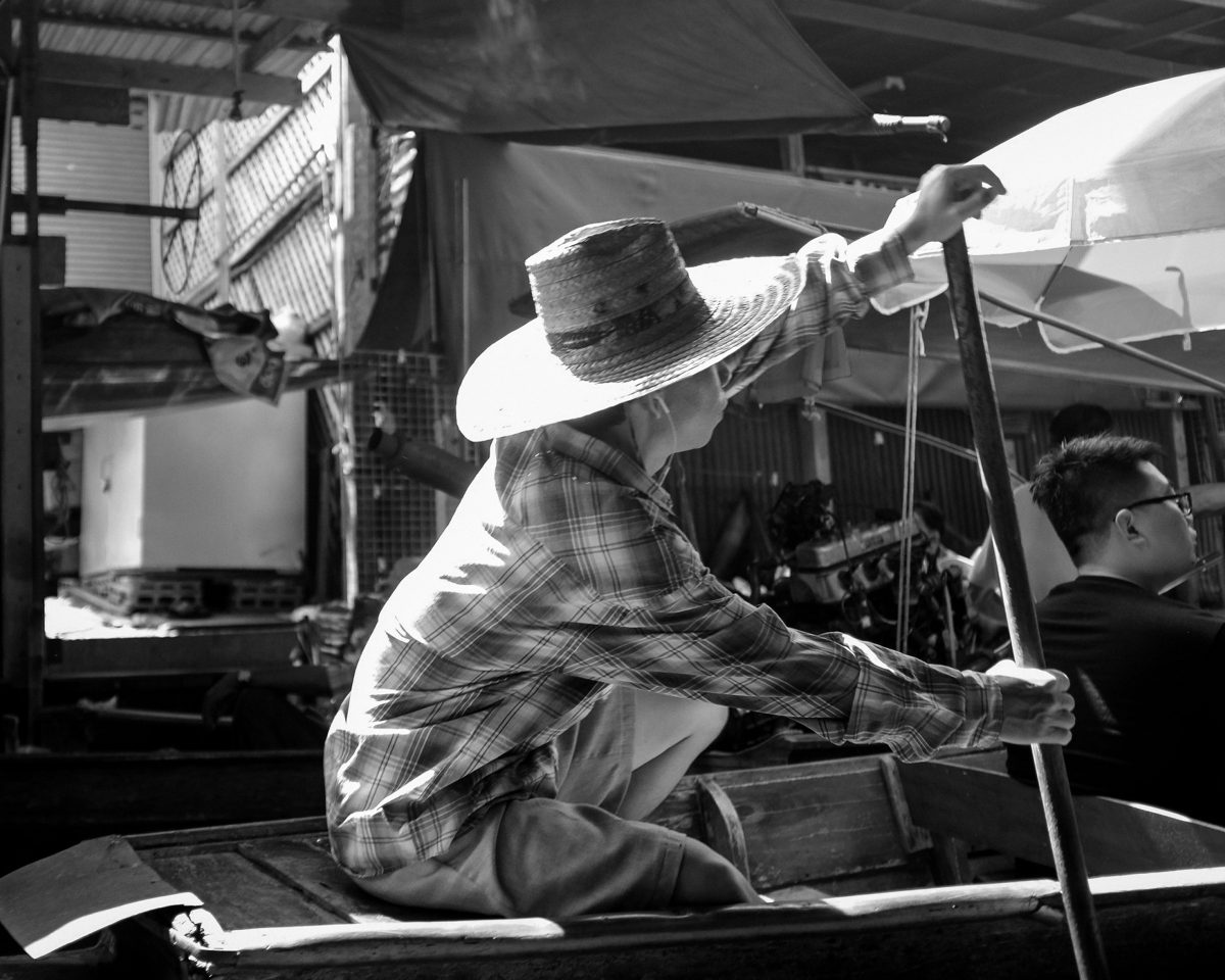 A boatman in a wide-brimmed hat leans on his pole at the back of a long-tail boat