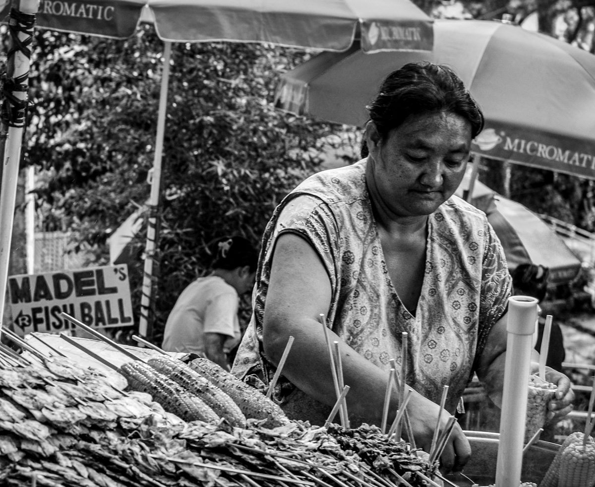 A woman grills skewers at a fish-ball stall under market umbrellas