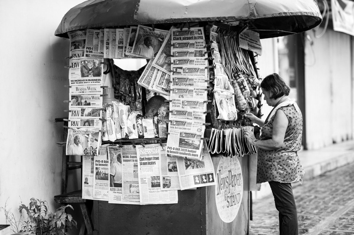 A vendor stands beside a covered newsstand wall-to-wall with newspapers and magazines