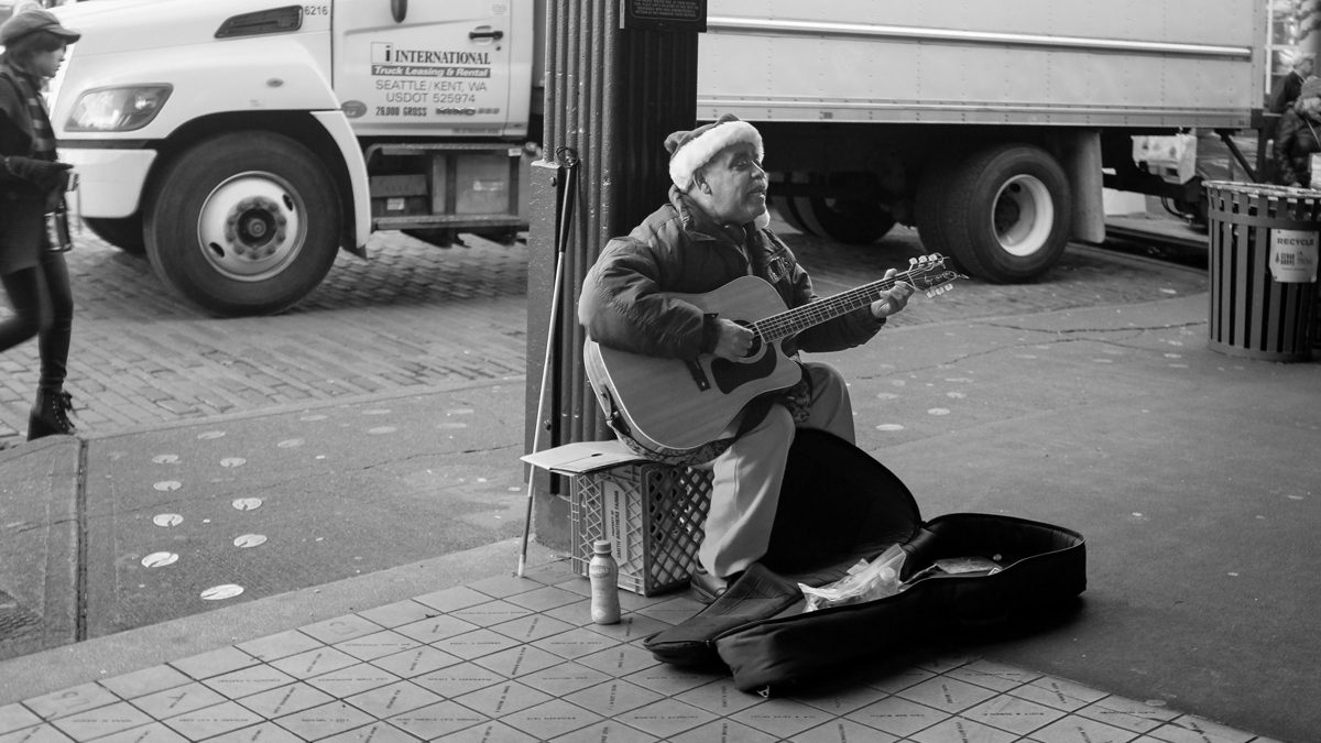 A busker in a woolly hat plays acoustic guitar on a paved corner beside his open guitar case