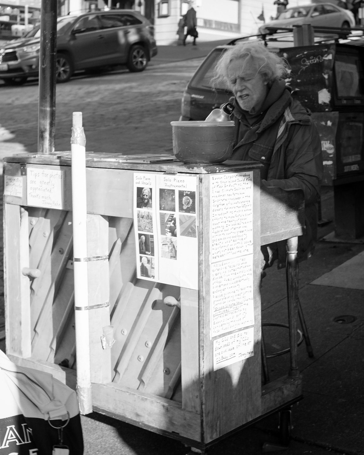 An older man sits at a battered street piano covered in notices