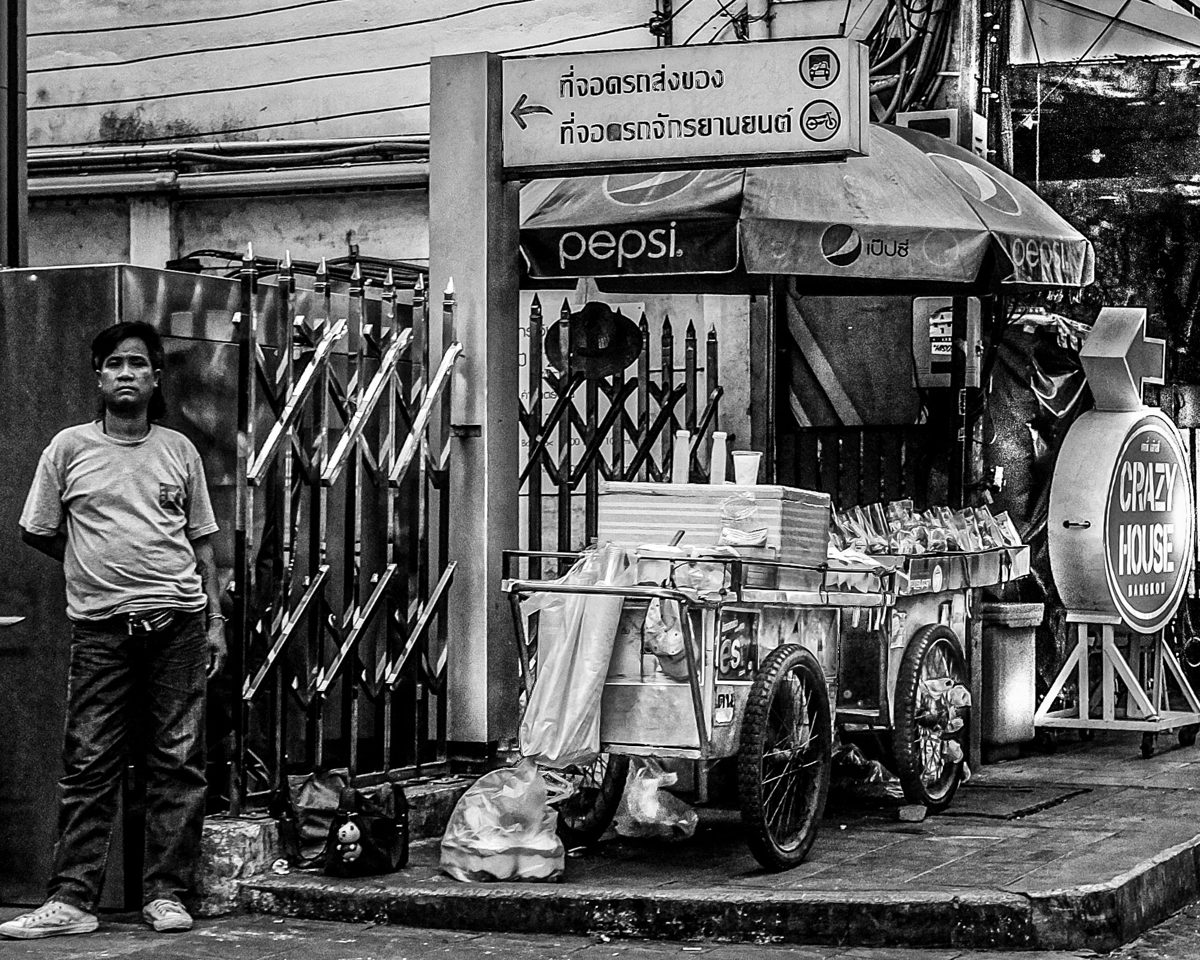 A man in a T-shirt stands beside a loaded food cart and a drinks stall at the side of a road