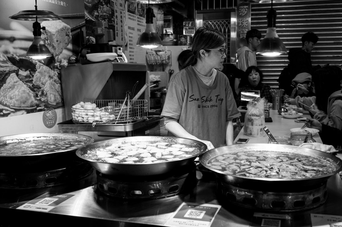 A cook at a row of glowing woks stirs a dish in a busy night kitchen